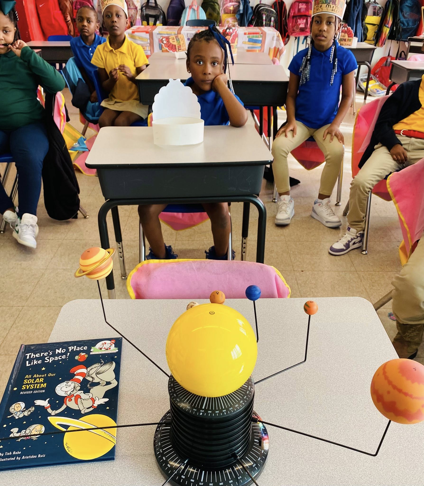 A Dr. Seuss book and a planet model on a desk with kids in the background.